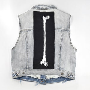Op de afbeelding: Een lichtblauw denim vest met een zwarte patch op de rug met een witte omtrek van een menselijk dijbeen.