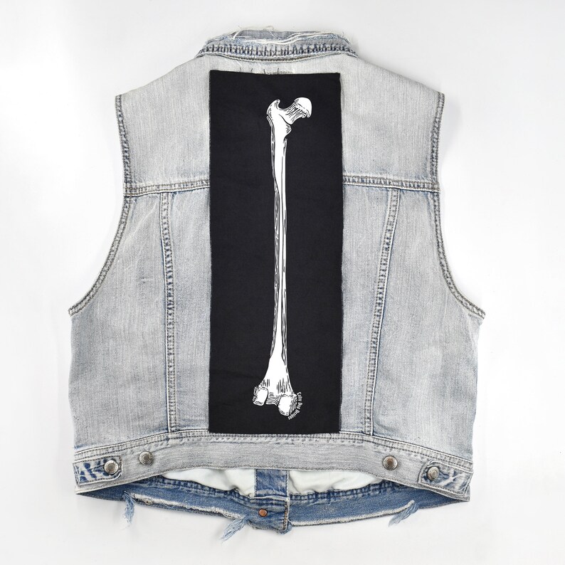 Op de afbeelding: Een lichtblauw denim vest met een zwarte patch op de rug met een witte omtrek van een menselijk dijbeen.