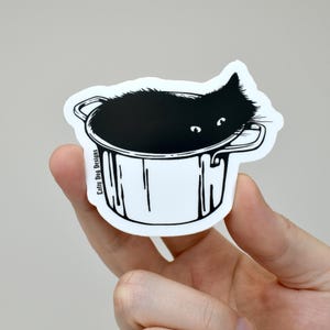 Puede incluir: Una pegatina troquelada que muestra a un gato negro asomándose desde el interior de una olla blanca con detalles de líneas negras. El gato tiene ojos blancos visibles y el texto Lofty Dog Designs está impreso verticalmente en el lado izquierdo.