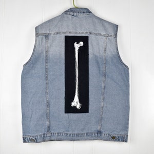 Op de afbeelding: Een lichtblauw denim vest met een zwarte patch op de rug met een wit skeletbeen.