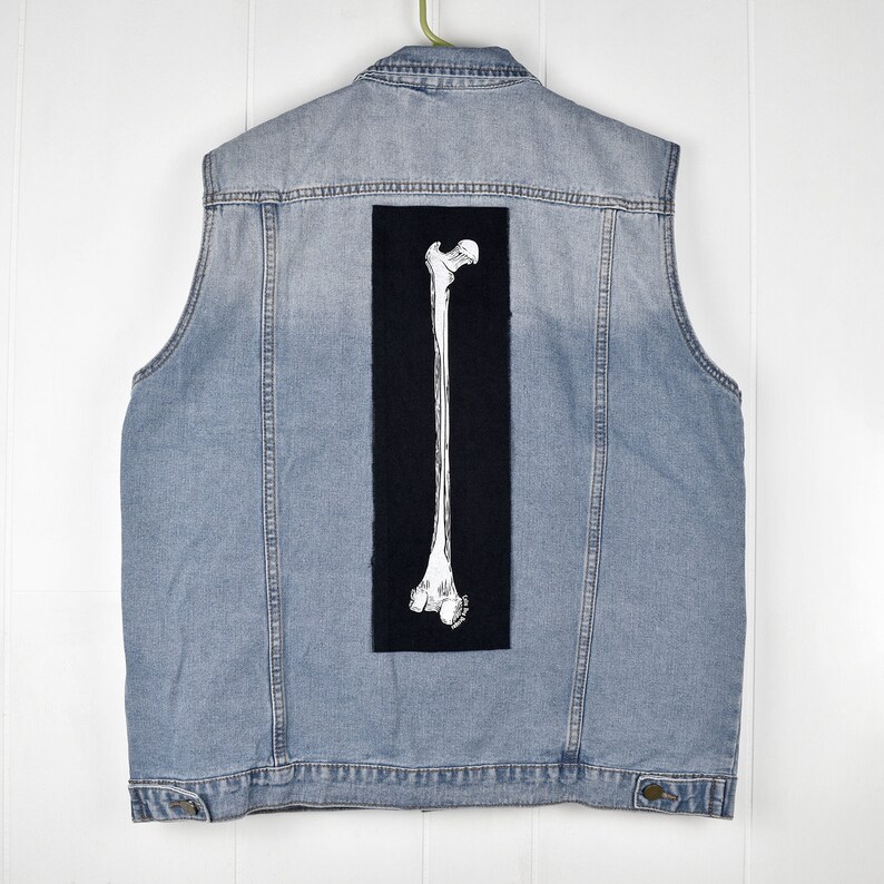 Op de afbeelding: Een lichtblauw denim vest met een zwarte patch op de rug met een wit skeletbeen.