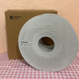 May include: A white PLA Basic 3D printer filament spool with a perforated design. The spool is next to a brown cardboard box with the Bambu Lab logo. The text "PLA Basic" is visible on the spool.