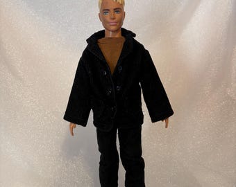 May include: A doll wearing a black corduroy jacket and matching pants over a brown sweater. The doll has short, blonde hair and brown shoes. The jacket has three buttons and a collar.