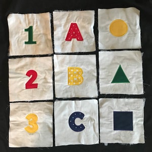 May include: Nine white squares with fabric appliques. The squares are arranged in a 3x3 grid. The top row features the number 1 in green, the letter A in red, and a yellow circle. The middle row features the number 2 in red, the letter B in yellow with white polka dots, and a green triangle. The bottom row features the number 3 in yellow, the letter C in blue with white polka dots, and a dark blue square.