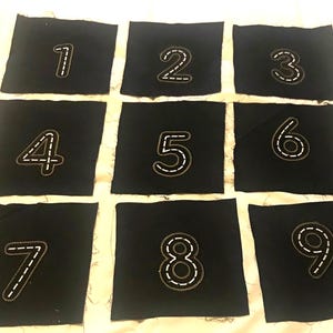 May include: Nine black fabric squares with white stitching, each featuring a number from 1 to 9.