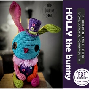 Peut inclure: Un lapin crocheté coloré nommé Holly, avec un corps bleu, des oreilles roses et vertes, et un chapeau haut de forme violet. Le lapin a des yeux en boutons et un nez en forme de cœur. Le texte indique "HOLLY the bunny" et "PDF Crochet Pattern".