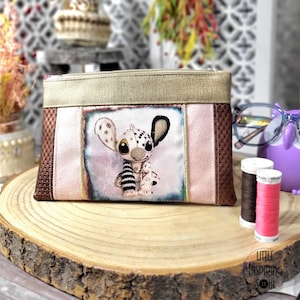 Peut inclure: Une pochette zippée faite à la main avec un personnage de dessin animé. La pochette a un dessus doré, un centre rose et des côtés marron. Le personnage a de grandes oreilles et un motif dalmatien. Le texte "Little Inspiring Soul" est visible.