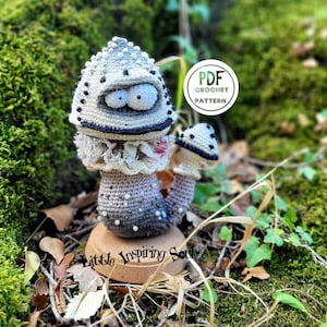 Peut inclure: Un champignon crochet fantaisiste avec un chapeau moucheté gris et blanc, des détails noirs et de grands yeux globuleux. Il possède un col à volants et une tige dégradée, posé sur une petite base en bois. Le texte "Little Inspiring Soul" est visible. Logo "PDF Crochet Pattern".