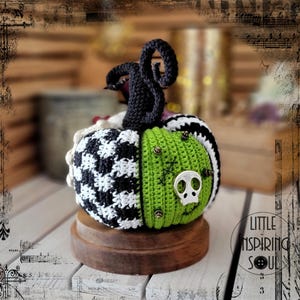 May include: A crocheted pumpkin with a black and white checked pattern, green fabric, and a silver skull accent. The pumpkin is sitting on a wooden base.