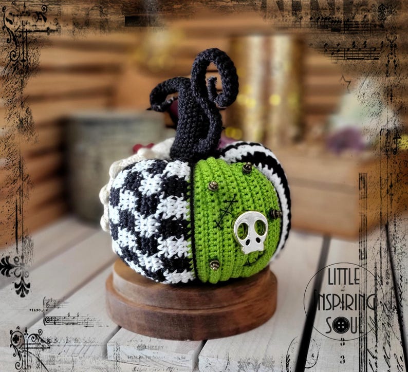 May include: A crocheted pumpkin with a black and white checked pattern, green fabric, and a silver skull accent. The pumpkin is sitting on a wooden base.