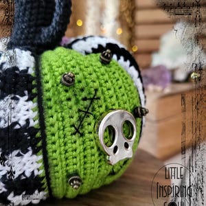 May include: A green crocheted pumpkin with black and white accents. The pumpkin has a skull-shaped metal embellishment and several smaller metal embellishments.