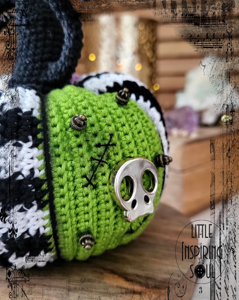 May include: A green crocheted pumpkin with black and white accents. The pumpkin has a skull-shaped metal embellishment and several smaller metal embellishments.