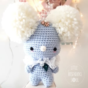Peut inclure: Un animal en peluche crocheté bleu clair avec des oreilles en pompon blanc, un col en dentelle blanche et un nœud vert. L'animal en peluche a des yeux noirs et un nez blanc. L'animal en peluche est assis sur un tabouret blanc. Le texte "Little Inspiring Soul" est visible dans le coin inférieur droit de l'image.