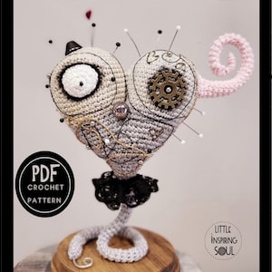 May include: A crocheted heart-shaped pincushion with a broken tail, a black lace trim, and a steampunk-inspired design. The pincushion is on a wooden base and has a pink tail.