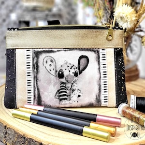 Peut inclure: Une pochette zippée noire et dorée avec un personnage de dessin animé en noir et blanc. La pochette a un dessus et une fermeture éclair dorés, avec un motif noir et blanc sur les côtés. Trois stylos à pointes dorées et roses sont devant la pochette.