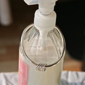 May include: A clear glass bottle with a white pump top and a silver metal ring around the bottle. The bottle is filled with a white liquid.