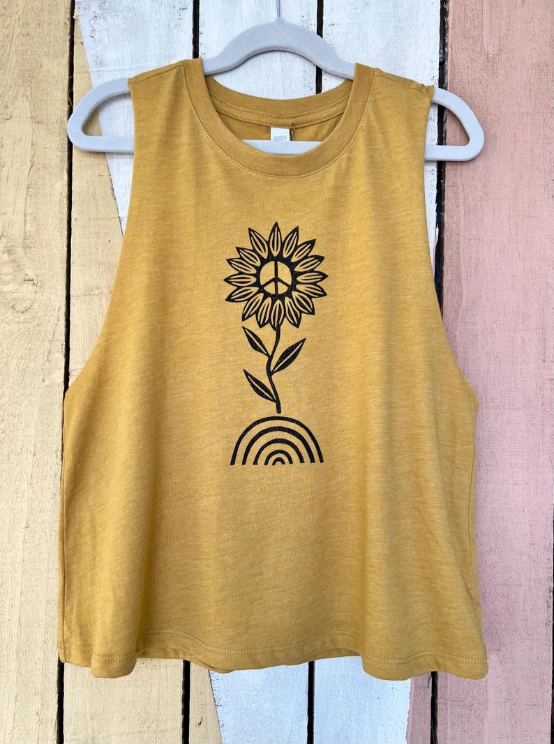 K&ouml;nnte beinhalten: Ein mussenfarbiges &auml;rmelloses Tanktop mit einem schwarzen Aufdruck einer Sonnenblume mit einem Peace-Zeichen in der Mitte. Die Sonnenblume w&auml;chst aus einem Regenbogen.