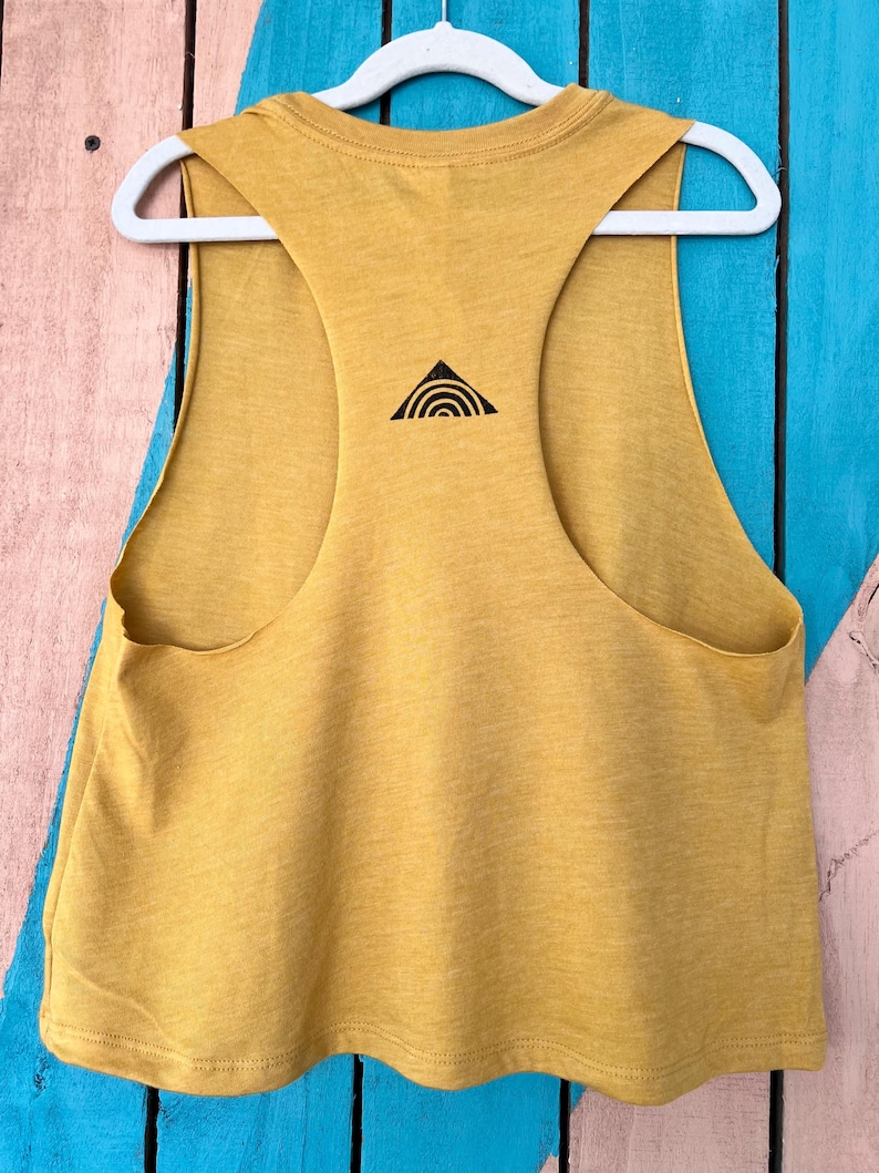 K&ouml;nnte beinhalten: Ein &auml;rmelloses Crop-Top in Senfgelb mit einem schwarzen Dreiecksdesign auf der R&uuml;ckseite. Das Dreieck hat eine geschwungene Linie im Inneren.