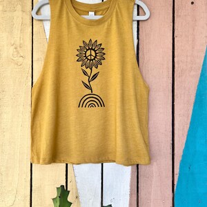 K&ouml;nnte beinhalten: Ein mussenfarbiges &auml;rmelloses Tanktop mit einem schwarzen Peace-Zeichen-Sonnenblumen-Motiv. Die Sonnenblume hat ein Peace-Zeichen in der Mitte und w&auml;chst aus einem Regenbogen.
