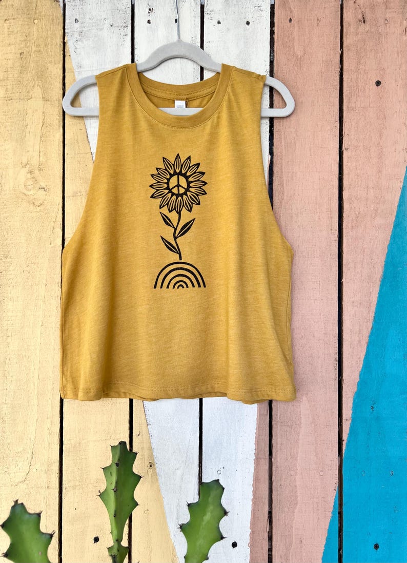 K&ouml;nnte beinhalten: Ein mussenfarbiges &auml;rmelloses Tanktop mit einem schwarzen Peace-Zeichen-Sonnenblumen-Motiv. Die Sonnenblume hat ein Peace-Zeichen in der Mitte und w&auml;chst aus einem Regenbogen.