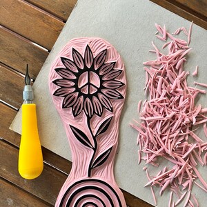 K&ouml;nnte beinhalten: Ein rosa Gummistempel mit einem schwarzen Design einer Sonnenblume mit einem Peace-Zeichen in der Mitte, einem Stiel und Bl&auml;ttern. Der Stempel hat auch ein schwarzes Regenbogen-Design am Boden.