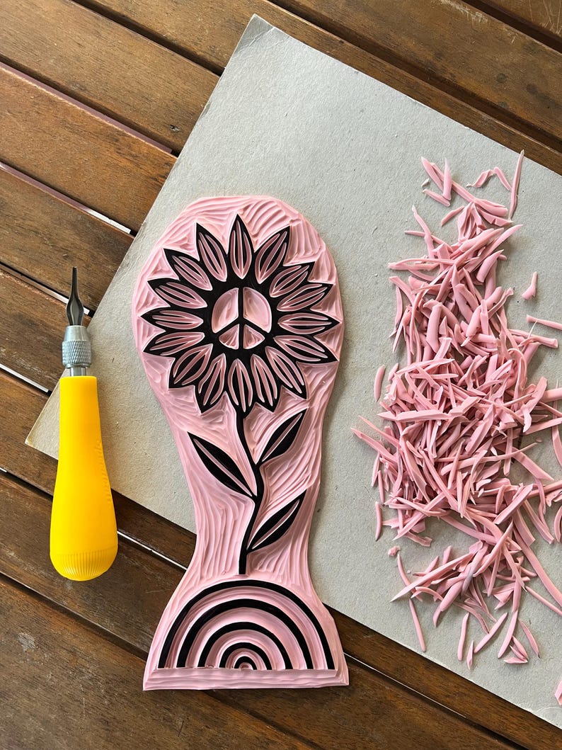 K&ouml;nnte beinhalten: Ein rosa Gummistempel mit einem schwarzen Design einer Sonnenblume mit einem Peace-Zeichen in der Mitte, einem Stiel und Bl&auml;ttern. Der Stempel hat auch ein schwarzes Regenbogen-Design am Boden.