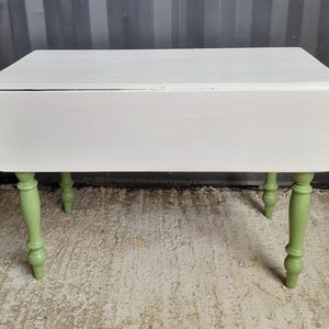 May include: A small, white drop-leaf table with four green, turned legs. The table has a rectangular top and is painted in a matte finish. The legs are a light green colour, contrasting with the white tabletop.