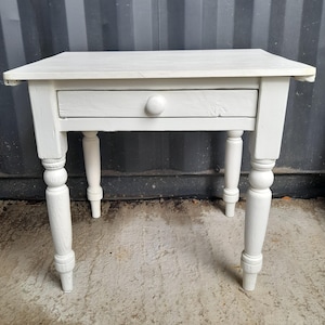 May include: A white painted wooden side table featuring a rectangular top, a single drawer with a round knob, and four turned legs. The table has a visible wood grain texture beneath the white paint finish and stands on four sturdy legs.