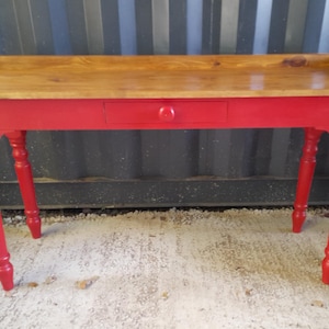 May include: A red painted wooden console table with a natural wood top. The table has a single drawer with a round knob and turned legs. The top has a raised edge on one side. The table is approximately 122 cm long.
