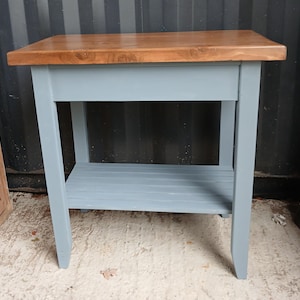 May include: A rectangular side table featuring a smooth, medium-brown wooden top and a light blue painted base. The table is constructed with four straight legs and includes a slatted lower shelf for storage, providing a stable surface area.