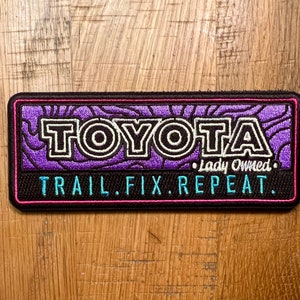 May include: Purple and black embroidered patch with the text "TOYOTA" in white and black lettering. The text "Lady Owned" is in white lettering below the word "TOYOTA". The text "TRAIL.FIX.REPEAT." is in blue lettering below the word "Lady Owned".