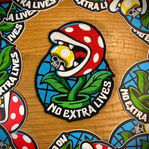 May include: Embroidered patch featuring a red and white spotted Piranha Plant with a skull-shaped mushroom in its mouth. The patch has a blue background and green leaves, with the text "NO EXTRA LIVES".
