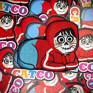 May include: Embroidered patch featuring a cartoon character in a red hooded jacket with a skull face. The character has a blue back and is surrounded by other similar patches. The word "CATCO" is visible.