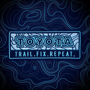 May include: Embroidered patch with the word "TOYOTA" in white and light blue, above the text "TRAIL. FIX. REPEAT." on a navy blue background. The background features a topographic map design in light blue.