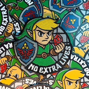 May include: Embroidered patch featuring a cartoon character in green and yellow, holding a red gem and shield. The patch has the text "NO EXTRA LIVES" in white. The background is filled with similar patches.