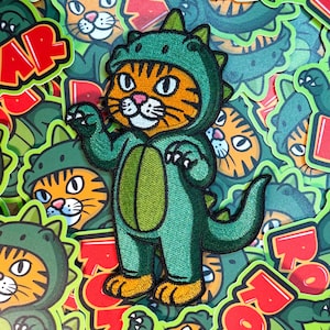 May include: Embroidered patch featuring an orange tabby cat wearing a green dinosaur costume. The patch has a detailed design with a hood, claws, and tail. The word "ROAR" is visible in red letters. Other patches with similar designs are in the background.