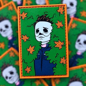 May include: A rectangular patch featuring a cartoon illustration of a character with a white mask and dark hair wearing a blue shirt. The character is surrounded by green foliage and orange maple leaves, all framed by a bright orange stitched border.