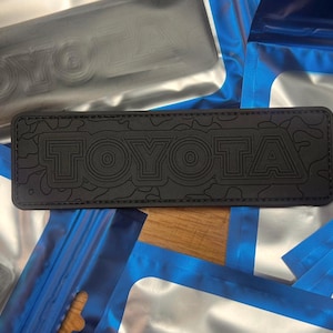 May include: Black leather patch with a camouflage pattern and the word "TOYOTA" embossed in white.