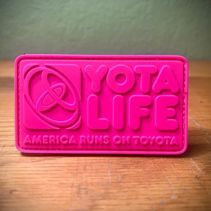 May include: A pink patch with the text "YOTA LIFE" and the Toyota logo. The text "AMERICA RUNS ON TOYOTA" is below the logo.