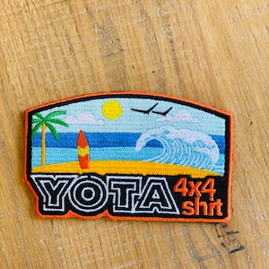 May include: An orange, black, and white embroidered patch with a beach scene, palm tree, surfboard, and wave. The text "YOTA 4x4 shirt" is on the patch.