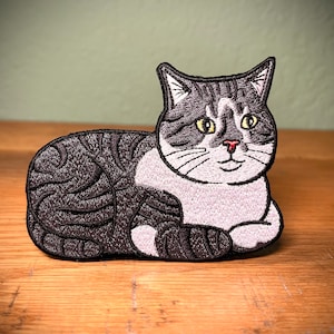 May include: A black and white embroidered patch of a cat lying down. The cat has a white chest and paws, and its eyes are yellow.