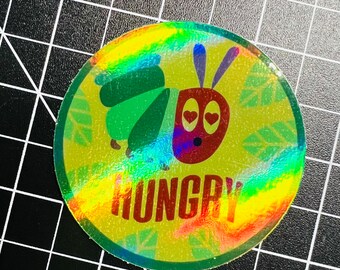 May include: A round, iridescent sticker with a green caterpillar and a red, cartoon caterpillar with googly eyes. The red caterpillar has the word "HUNGRY" written below it.