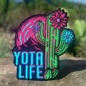 May include: A black embroidered patch with a green cactus, pink and blue flowers, a pink and blue sunset, and the text "YOTA LIFE".