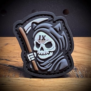 May include: A black patch featuring a grim reaper design. The reaper is depicted with a white skull, a gray hood, and a brown scythe. The Roman numeral "IX" is above the skull. The patch has a black border and is set against a wooden surface.