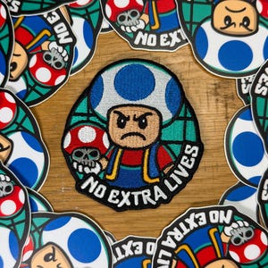 May include: Embroidered patch featuring a cartoon character with a blue mushroom cap and a stern expression, holding a skull-shaped mushroom. The patch has the text "NO EXTRA LIVES" and is surrounded by similar patches.