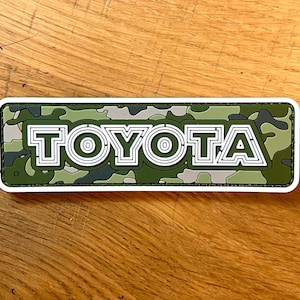 May include: A green and white camouflage patch with the word "TOYOTA" in white outlined letters.
