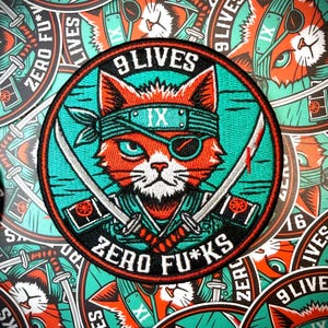 May include: Embroidered patch featuring a red fox with a teal headband and eye patch, holding two crossed swords. The patch reads "9 LIVES" and "ZERO FU*KS" in white lettering, set against a teal and black background.