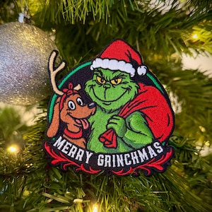 May include: Embroidered patch featuring the Grinch in a Santa hat and a dog with antlers. The Grinch is green and holding a red sack. The patch has the words "MERRY GRINCHMAS" in white lettering on a red banner.