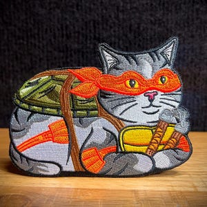 May include: A gray cat wearing an orange and red ninja turtle mask and a green shell on its back. The cat is holding a pair of katanas in its paws.