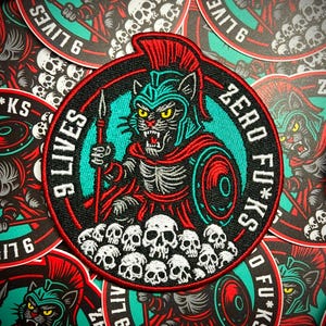 May include: Embroidered patch featuring a fierce cat warrior in a helmet, holding a spear and shield, surrounded by skulls. The patch has the text "9 LIVES ZERO F*CKS" in a circular design, with red, black, teal, and white colors.